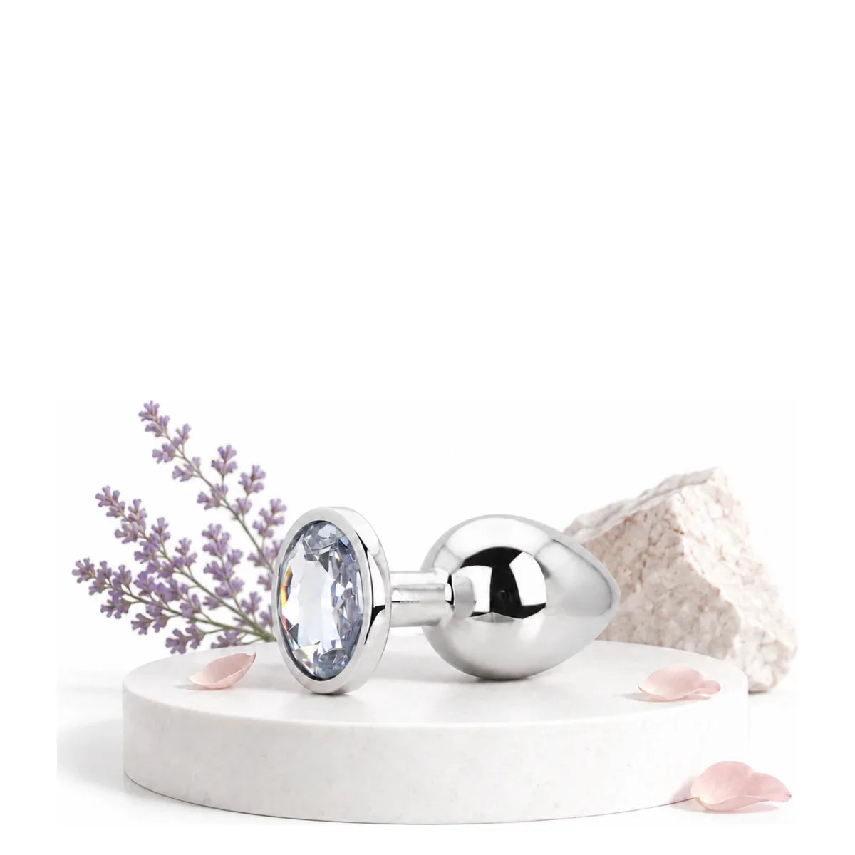 Silberfarbener Analplug mit facettiertem Schmuckstein auf einem weißen Podest neben Lavendel und Rosenblättern.