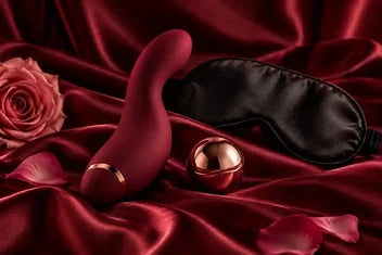 Weinroter Vibrator, schwarze Augenmaske und roségoldfarbene Kugel auf Satin mit Rosenblättern.